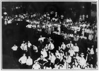 Black-and-white image of two crows of people, one sitting and standing in rows, the other standing in front of the first. White shirts stand out against a black background